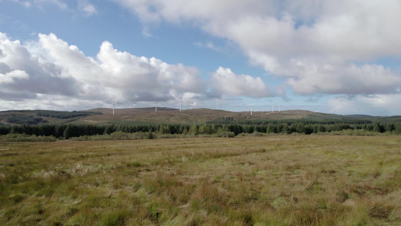 공중 드론 영상은 킨타이어 반도, 아가일, 스코틀랜드에 있는 상업용 침엽수의 임업 농장으로 둘러싸인 스코틀랜드 풍력 발전소의 풍력 터빈을 향해 들판 위로 빠르고 낮게 날아갑니다.