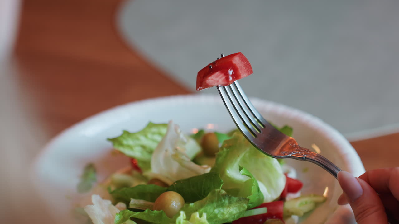 Fresh salad with fork lifting tomato, bowl of lettuce and olives on wooden table, closeup of hand picking cherry tomato, bright natural light, slow deliberate bite, healthy home dining mood, soft