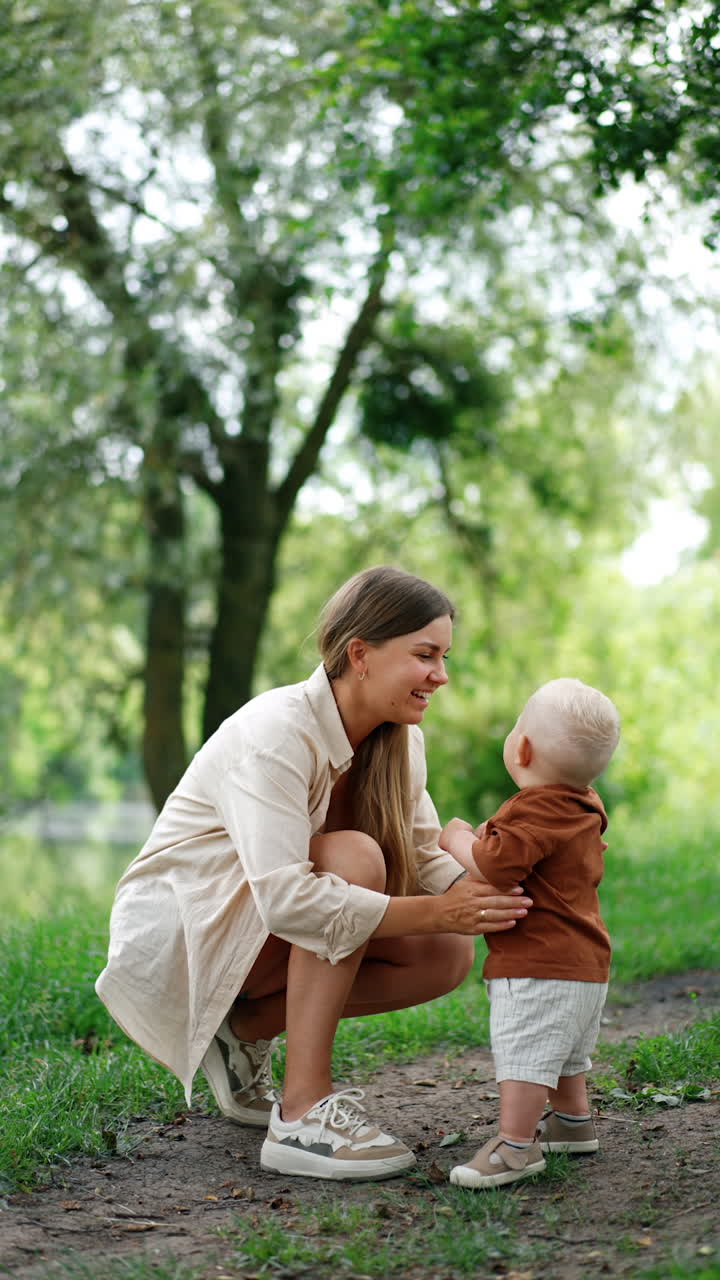 Mother sits squatted near her little son. Woman holds her lovely baby standing unsteadily. Vertical video.