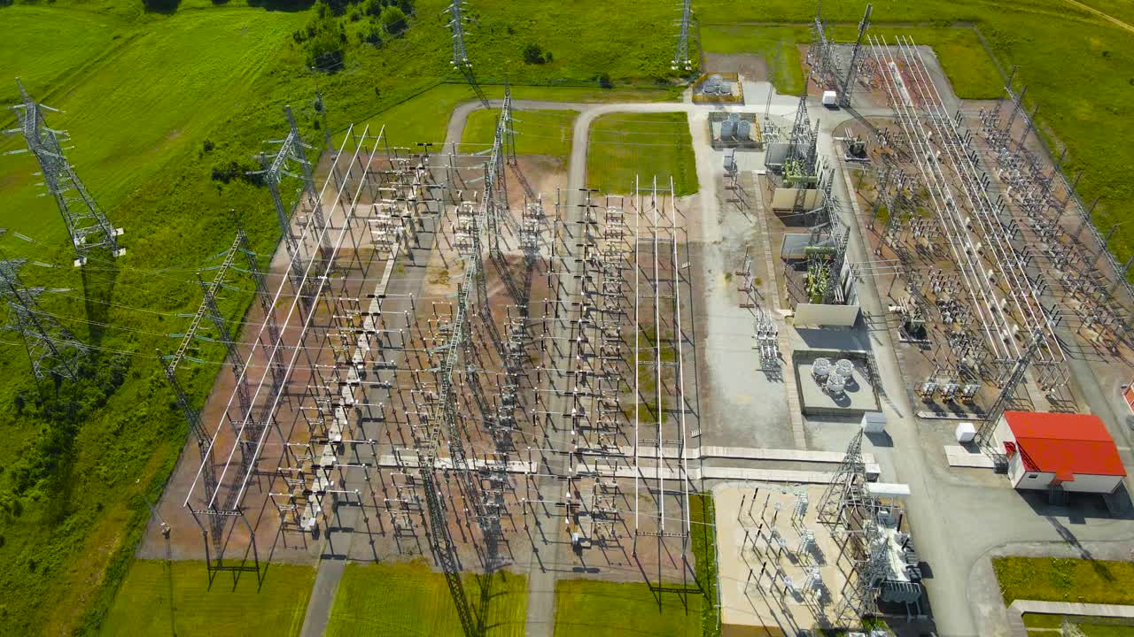 Aerial drone footage orbiting around a modern electrical substation power station during a sunny day in a rural countryside with high voltage and dangerous power cables and transformers, control room