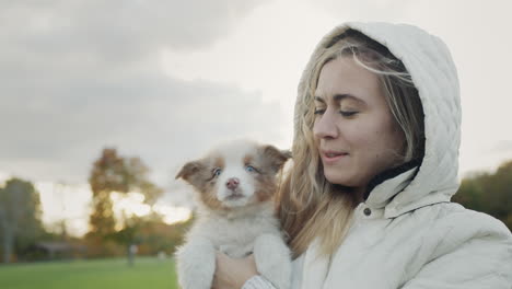 Una-Mujer-Joven-Sostiene-Un-Lindo-Cachorro-En-Sus-Manos,-Un-Cachorro-Le-Lame-La-Cara.-Caminando-Juntos-En-Un-Parque-De-Otoño.