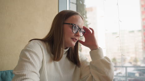woman seated near window leans back with gentle smile and closed eyes during moment of thoughtful mood swing surrounded by soft natural light and city view outside