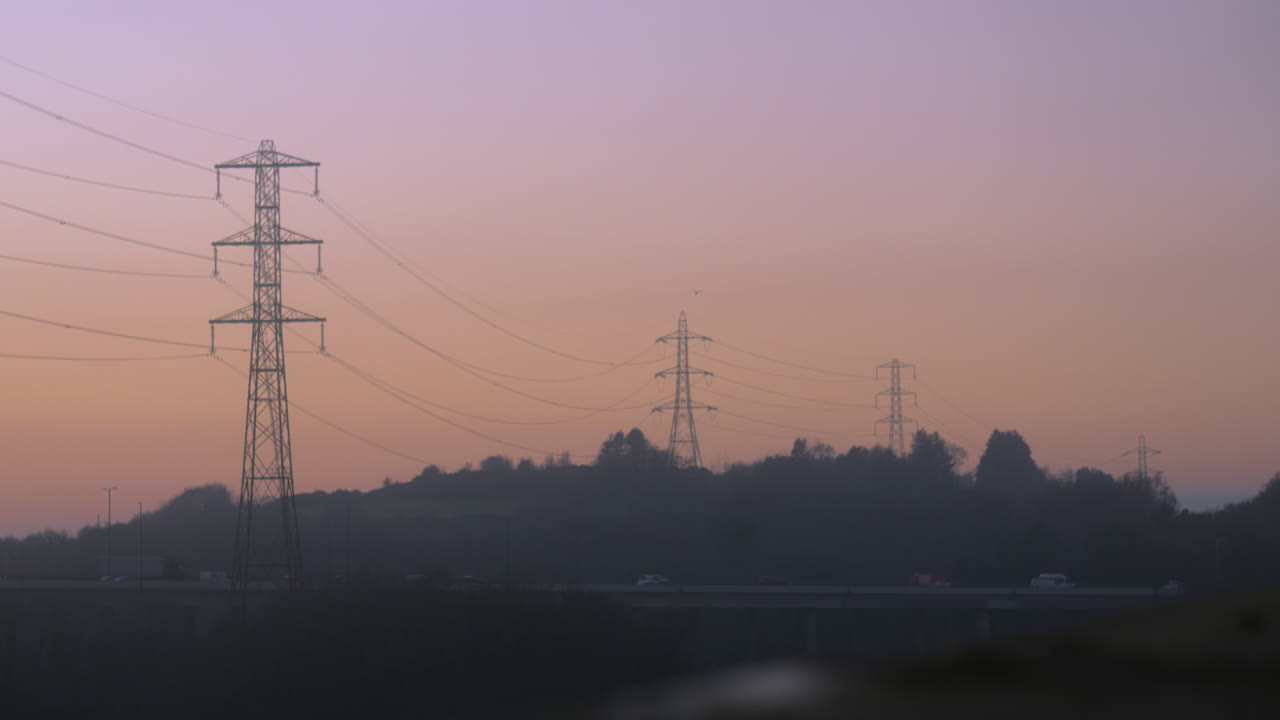 Electricity Pylons Carrying Energy Over Busy Motorway with Cars and Trucks Travelling Home During Sunset. M4 Road Bridge Near Swansea, Wales 4K.