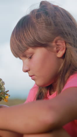 la niña mira pensativa a las flores en su mano mientras está sentada en el campo rural, la suave luz del sol destaca su expresión mientras admira los delicados pétalos con vastas tierras de cultivo
