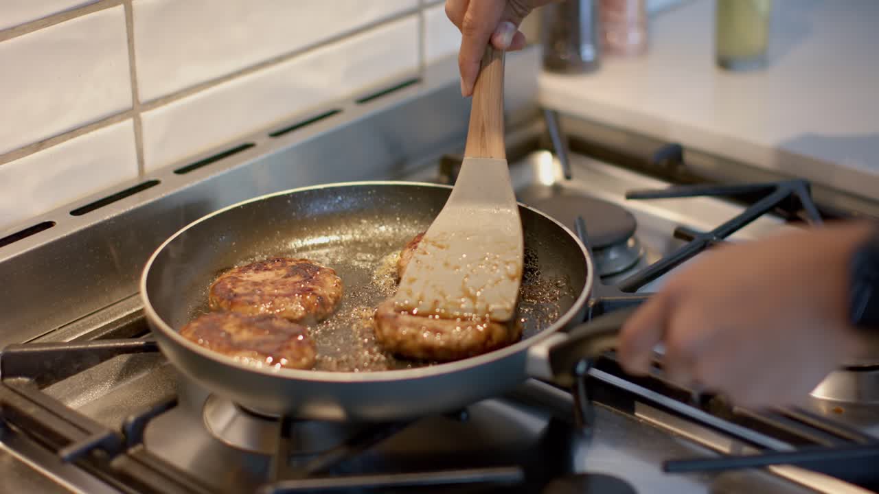 Padella sul fuoco che sfrigola con hamburger di manzo; il cuoco li gira e li bagna con una spatola per sigillarli.