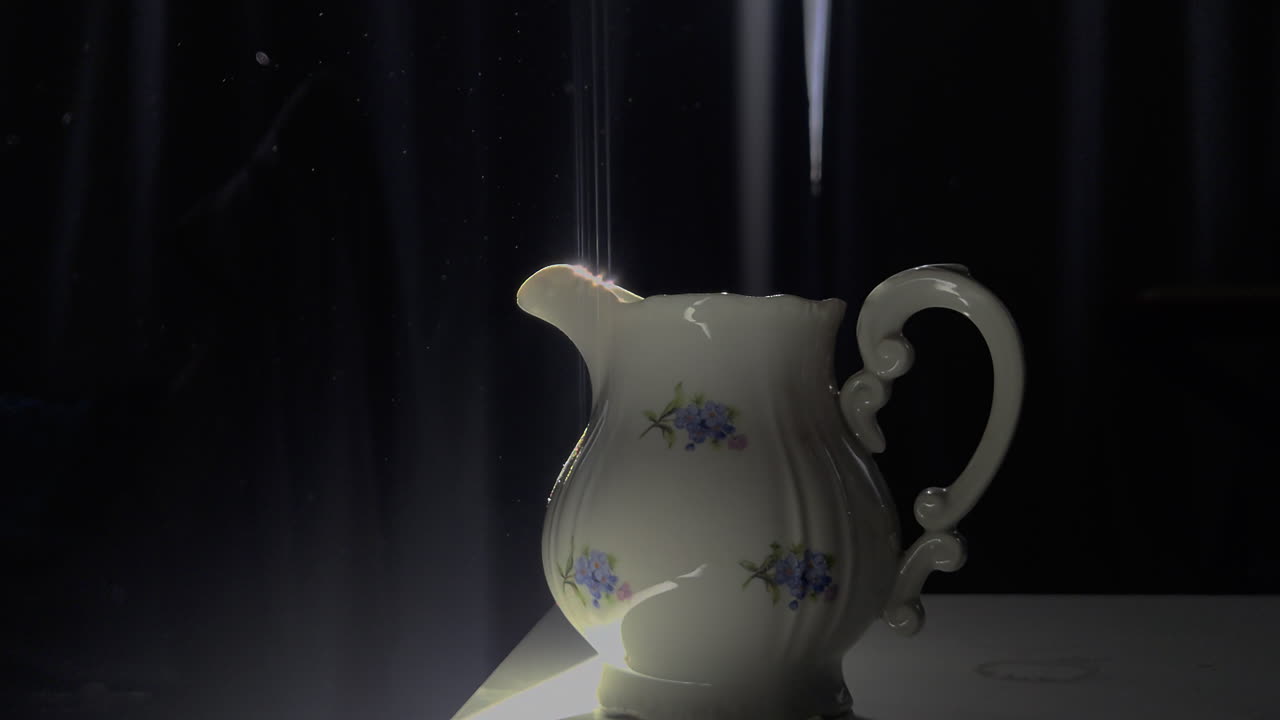 Un'inquadratura statica e suggestiva cattura una piccola lattiera in ceramica con motivi floreali su un tavolo. Un fascio di luce intenso fende l'oscurità, illuminando particelle di polvere visibili che danzano nell'aria soprastante.