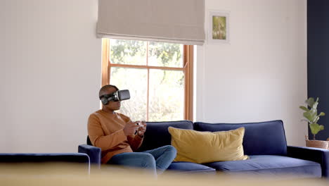 african american man sitting on sofa using vr headset and playing video games at home, slow motion