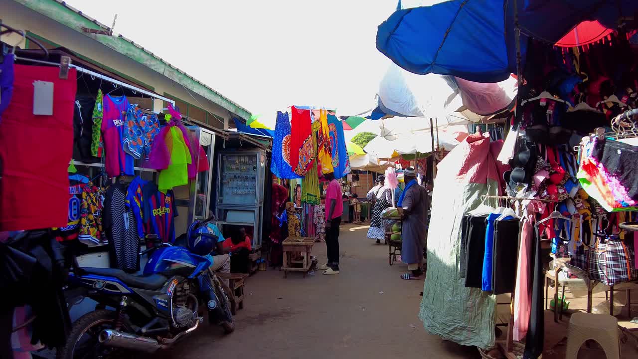 POV walk through bustling Serekunda Market with diverse goods, vendors, and crowds as people carry boxes down narrow hallways