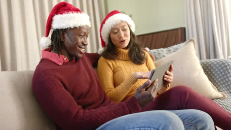 happy diverse couple in christmas hats using tablet on couch at home, slow motion