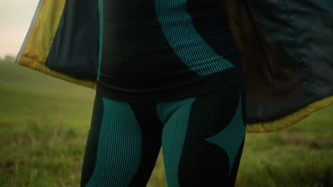 close-up of individual in green and black suit unzipping yellow jacket while standing in vast open field with trees in the distance, under a cloudy sky