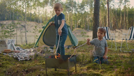 two boys sitting by a fire against a tent in the woods on the shore of the lake fry marshmallows on fire. brothers 3-6 years together burn sticks on fire