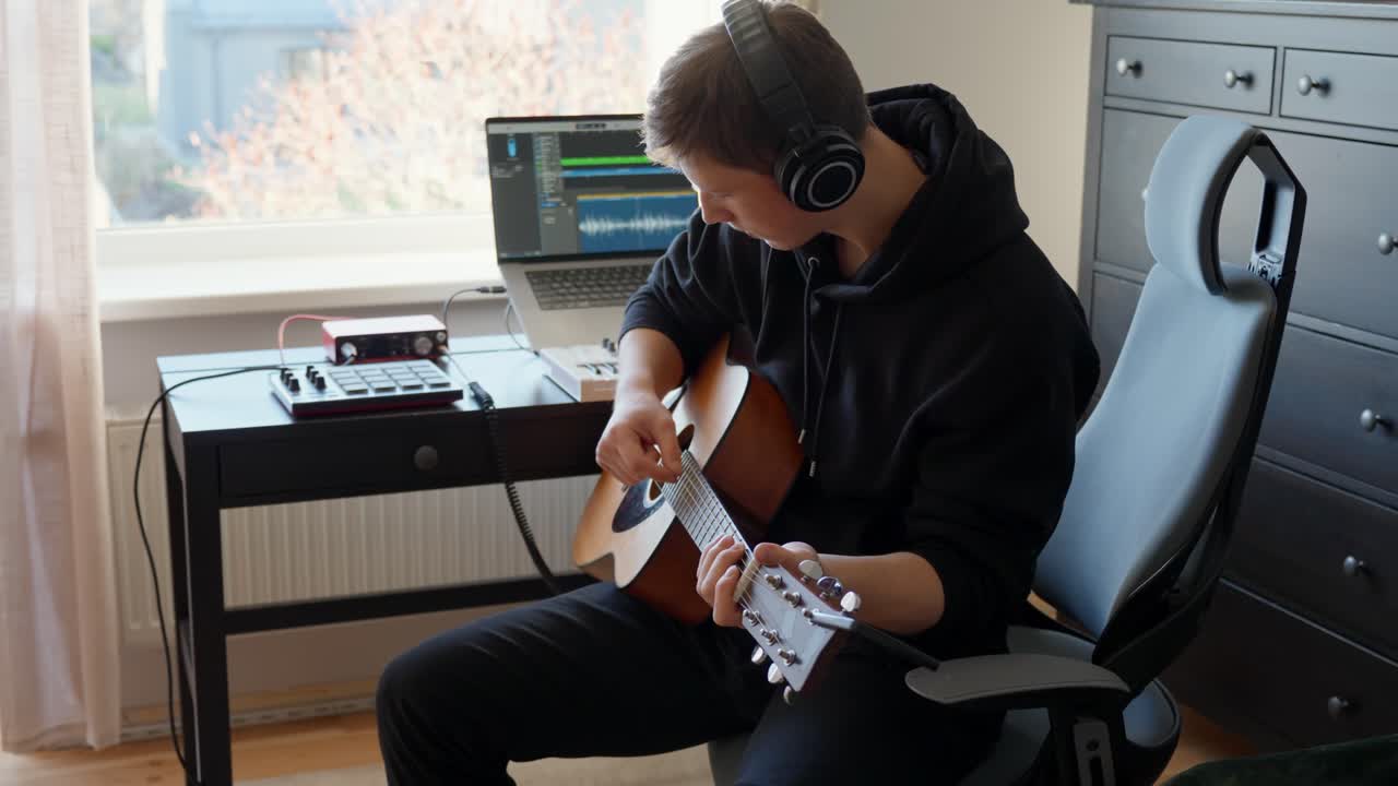 Young music producer wearing headphones creating a song playing acoustic guitar in a home studio, using a laptop with music software and recording equipment