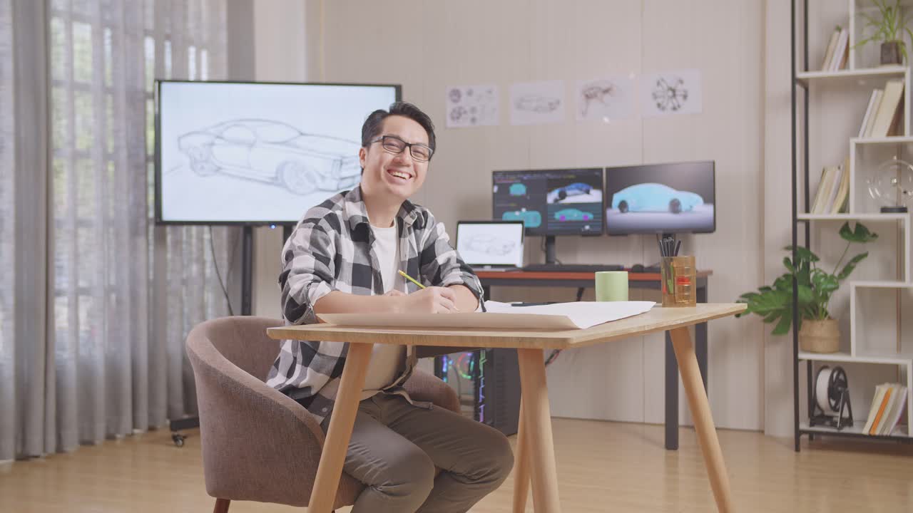 tv와 컴퓨터 디스플레이 3d 전기 자동차 모델과 함께 스튜디오에서 테이블에 자동차 디자인 스케치를 작업하는 동안 카메라에 미소 짓는 아시아 남성