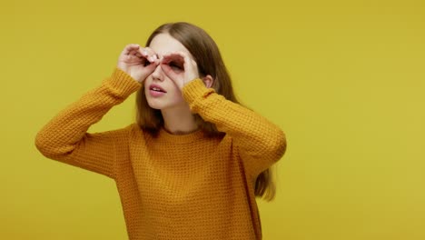 cute inquisitive girl in casual clothes looking around through fingers imitating binoculars, saying wow