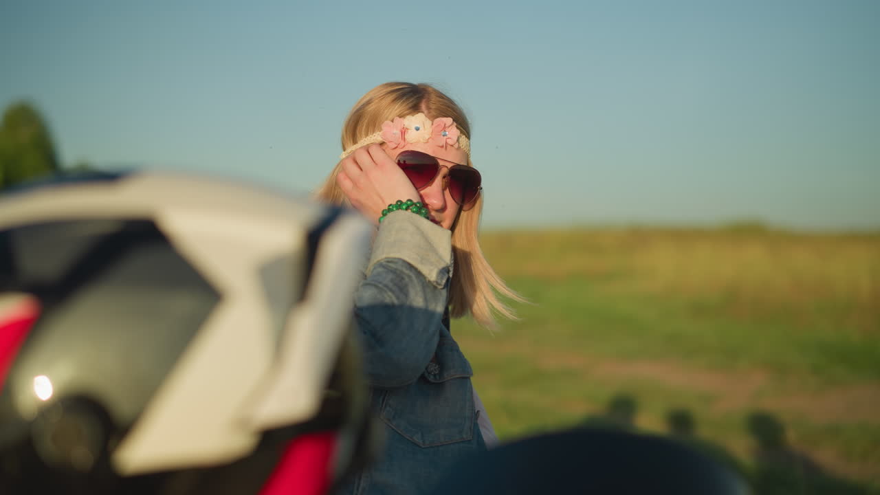 una mujer con una correa de flores y gafas de sol, con una pulsera de cuentas verdes en su muñeca, toca suavemente su cabello mientras descansa contra una motocicleta en un campo abierto