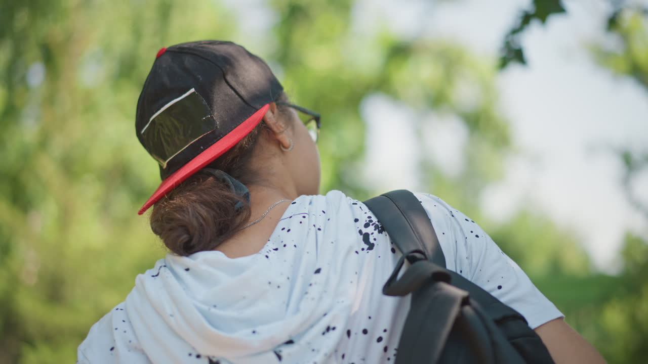 그늘진 길에서 모자를 거꾸로 눌러쓰고, 포니테일을 모자 아래로 넣고, 걷기 또는 하이킹을 준비하며 머리에 손을 대는 동작을 취하고, 캐주얼한 후드티를 입고 배낭끈을 맨 채 잎이 무성한 여름 풍경 속에 있는 아시아 젊은 성인