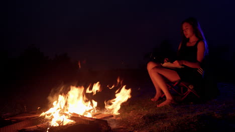 Hoguera-Acampando-Modelo-Femenino-Joven-Asiático-Sentado-Frente-Al-Fuego-Por-La-Noche-Mientras-Lee-Un-Libro