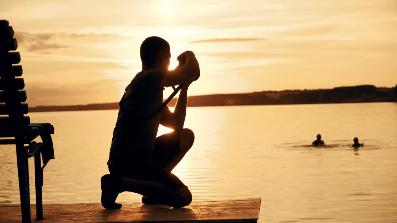 A man in shorts and a t-shirt is squatting and holding a camera focused on the sunset against the background of the forest and people in the river.