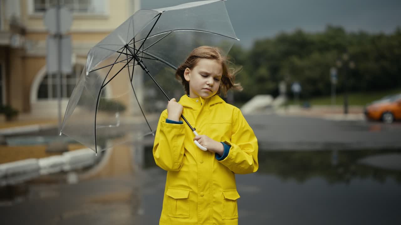 Sad teenage girl in a yellow jacket stands and holds an umbrella in her hands in a windy park after the rain