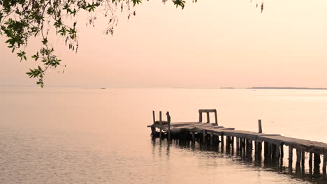 Alter-Holzsteg-Auf-Ruhigen-Wasserwellen-Auf-Dem-Meer-Mit-Wind,-Der-Grüne-Blätter-über-Einer-Kleinen-Insel-Bei-Sonnenuntergang-Umrahmt,-Bahrain