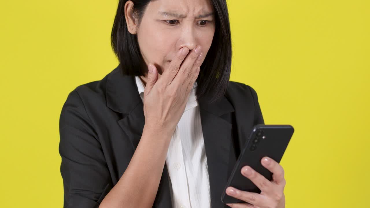 明るい黄色の背景の前に立つスーツ姿のプロフェッショナルな女性が、驚きの表情でスマートフォンのアラートを受け取ってショックを受けている様子。