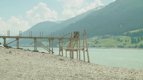 Wooden-Bridge-on-the-shore-of-Reschensee-in-Italy,-People-Kite-Boarding-on-a-Sunny-day-in-the-background