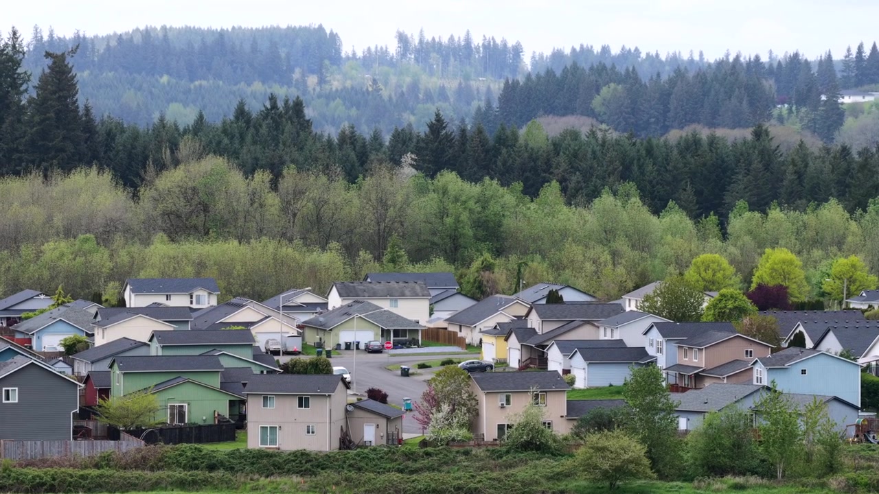 アメリカ、ワシントン州。鬱蒼とした丘の森に寄り添うように建つ2階建ての家とガレージ、緑の芝生、曲がりくねった道が広がる静かな郊外の住宅街を、曇り空の春の空の下で捉えた空撮ワイドショット。
