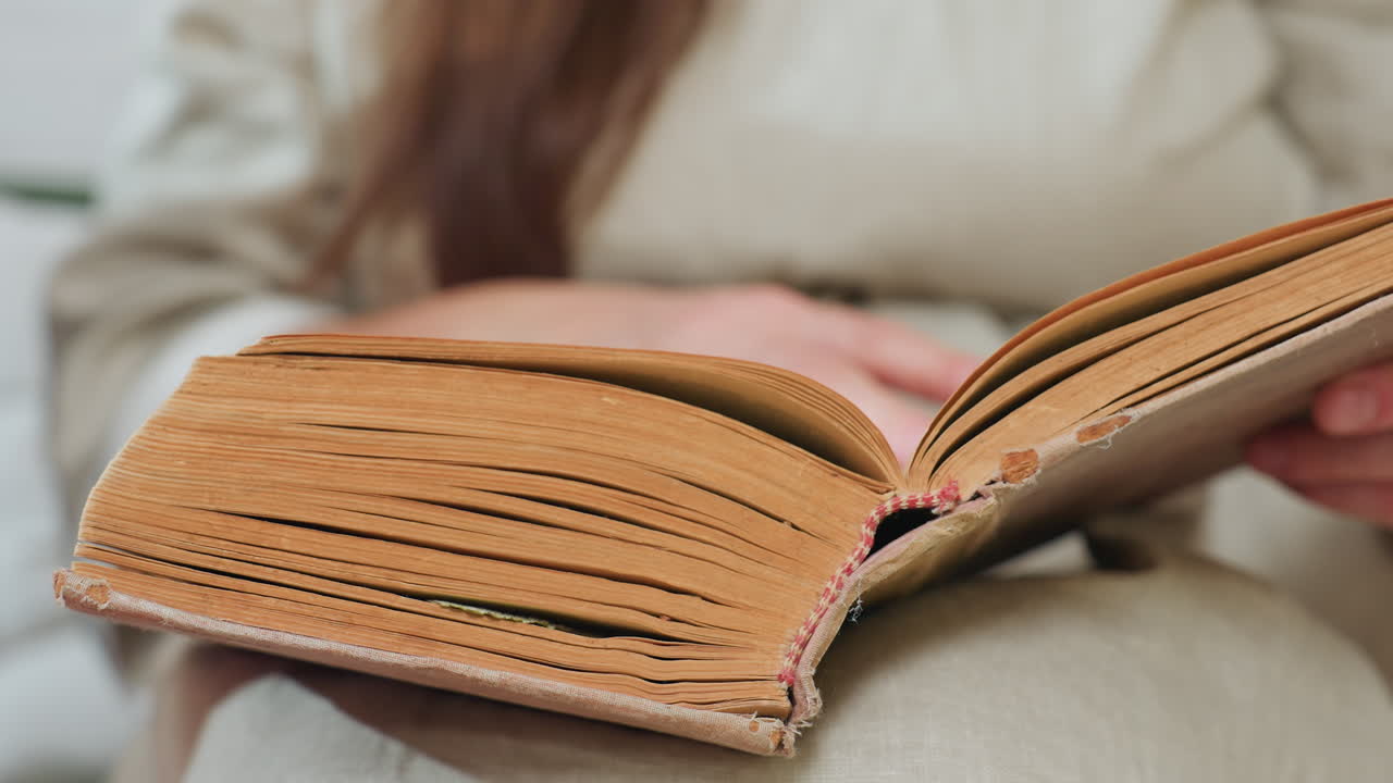 close view of old thick book with yellowed pages resting on fabric surface, blurred view of hand gently flipping through fragile pages, vintage atmosphere, cozy indoor setting, aged textured cover visible