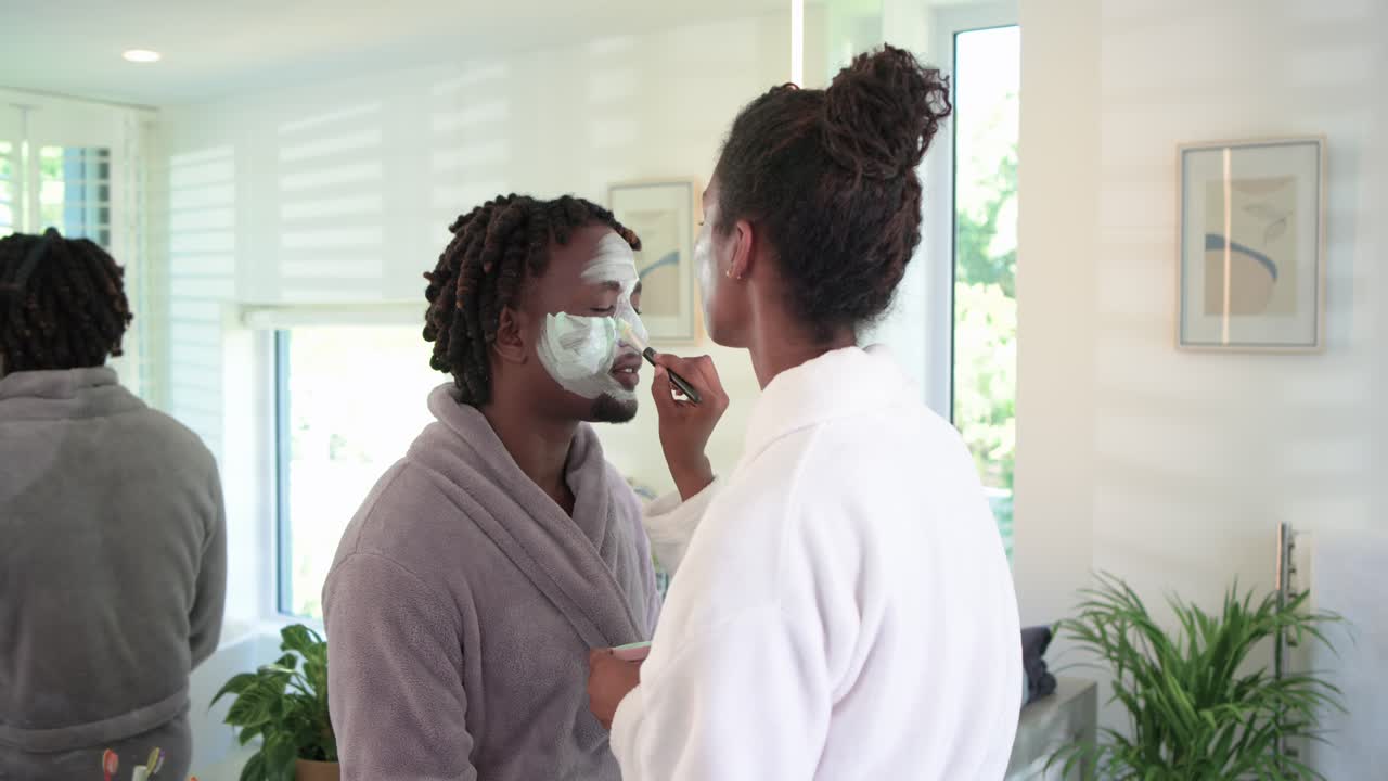 Coppia afroamericana in accappatoi per la cura della pelle, donna che intinge il pennello e applica una maschera all'uomo allo specchio.