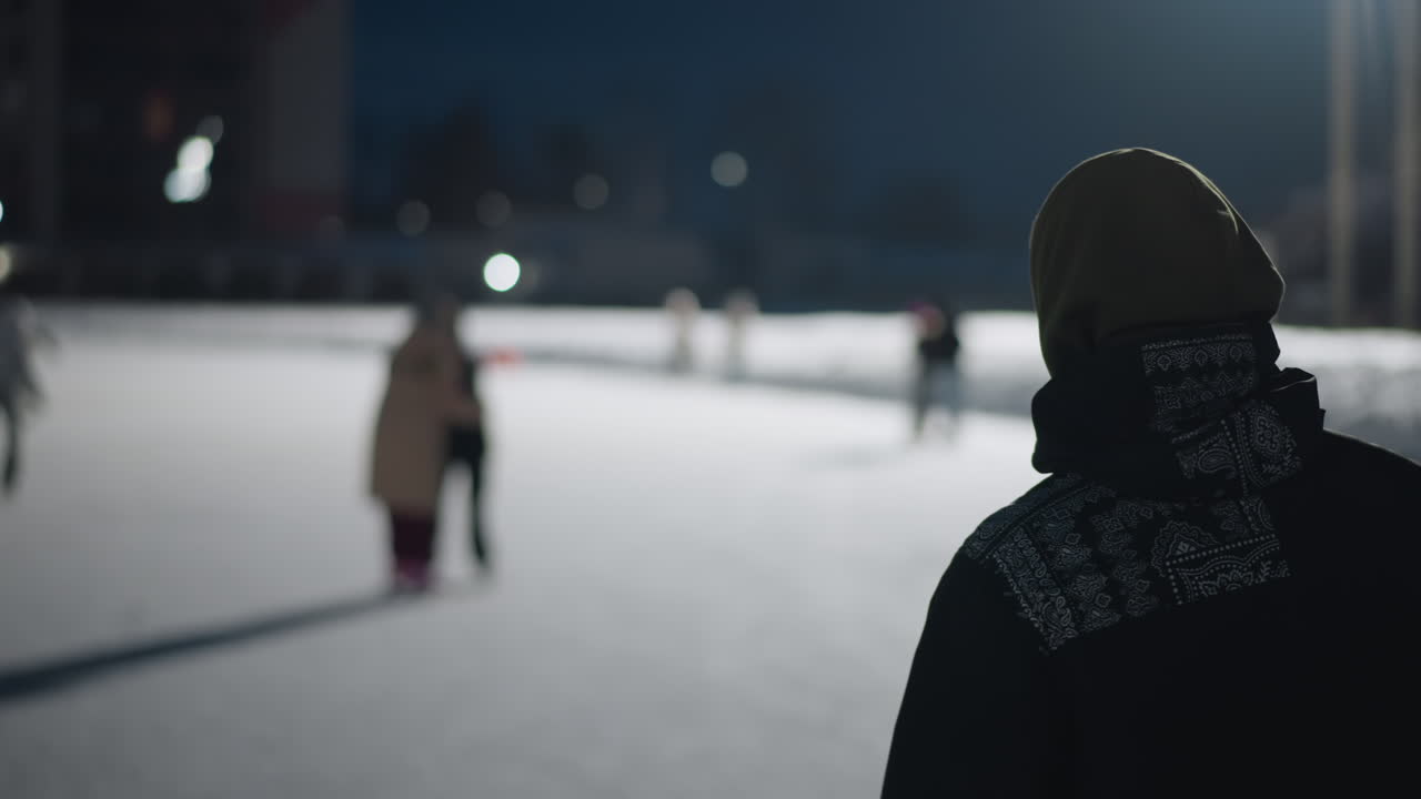 back view student skating on outdoor ice rink at night wearing hooded winter jacket cold breath visible in air blurred background of other skaters and glowing lights in frosty atmosphere