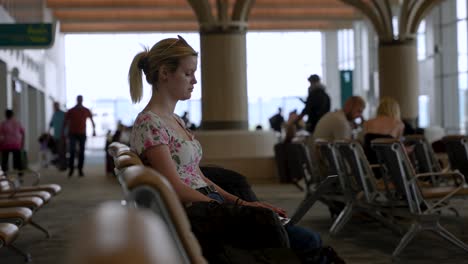 Mujer-Joven-Sentada-Sola-En-La-Terminal-Del-Aeropuerto-Usando-Una-Computadora-Portátil-Con-Pasajeros-Caminando-En-Segundo-Plano.