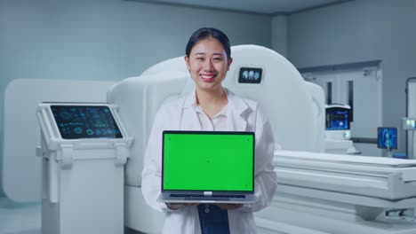 asian female doctor smiling and showing green screen laptop while standing with mri machine in the hospital