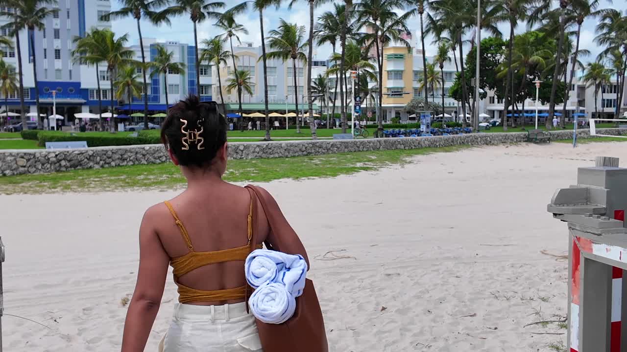 Carrellata di una ragazza turista asiatica che lascia la spiaggia a Miami, con hotel e appartamenti sullo sfondo - vista posteriore.