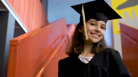 portrait of a happy preschool female student in cap and gown holding graduation diploma and looking at the camera