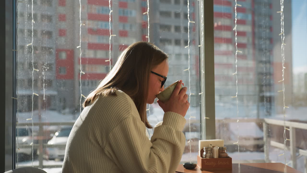 Side view of woman in cream sweater sipping coffee by large window, warm sunlight filling space, snowy city street and parked cars visible through glass, cozy and peaceful winter moment inside cafe