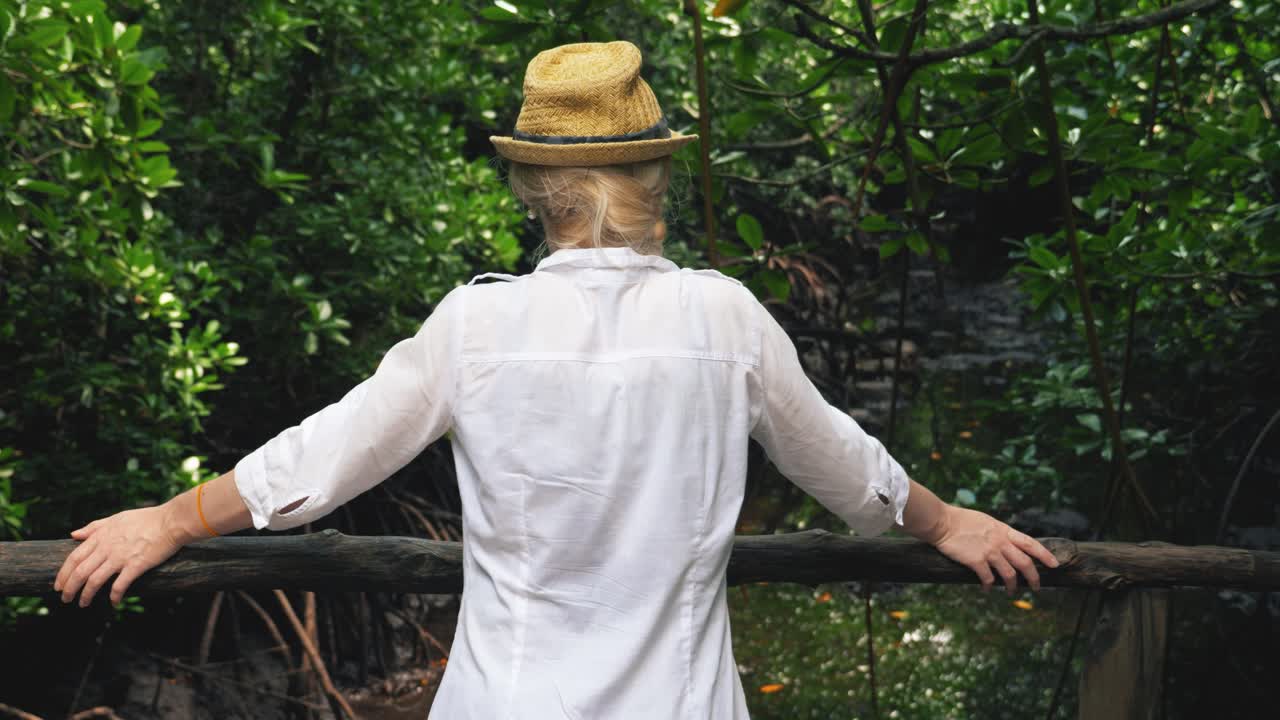 흰색 셔츠를 입은 어린 소녀가 오래된 목조 다리에서 녹색 빽빽한 정글을 바라보고 있습니다.