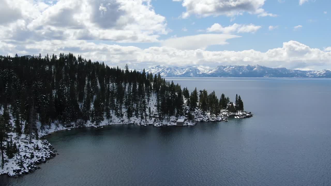A slow revealing 4K drone cinematic shot of the shoreline of Lake Tahoe on a sunny day. The winding coastline is filled with pine trees as you see the Sierra Nevada mountains of Nevada and California.