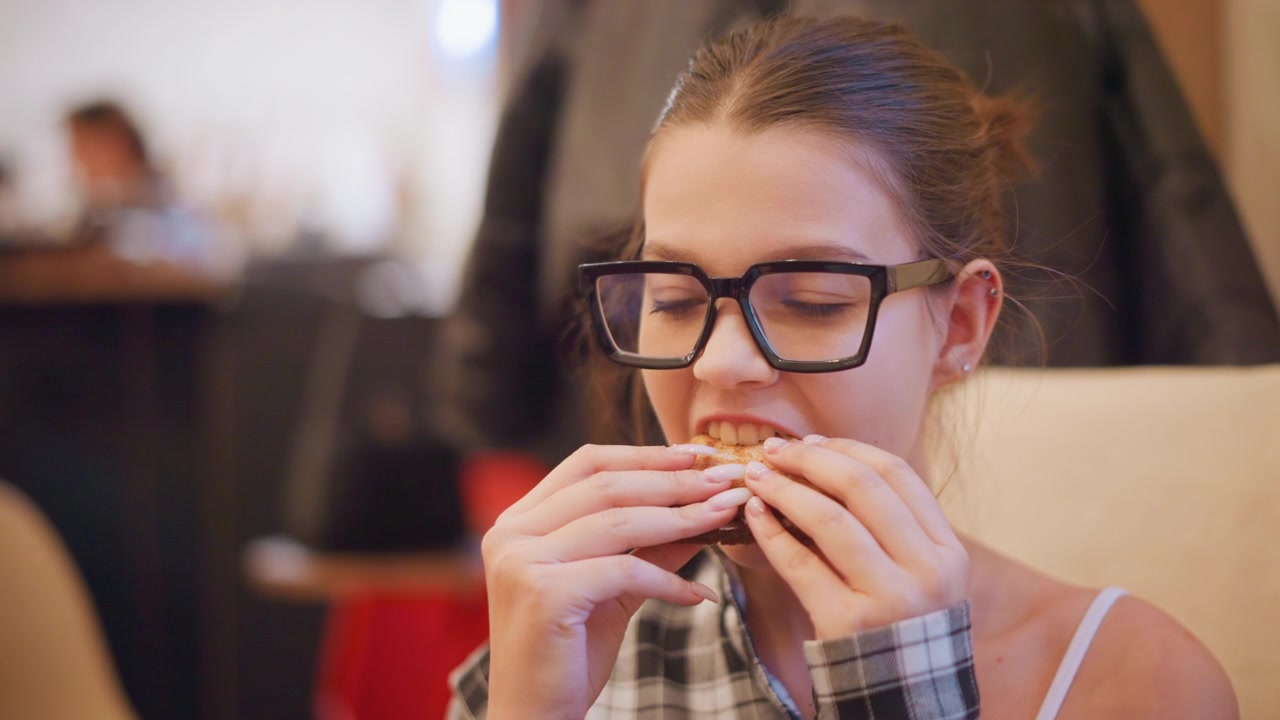 Close up of young woman eating food in indoor setting. Woman holds sandwich with both hands wearing glasses and casual clothing. background shows soft lighting and blurred figures suggesting