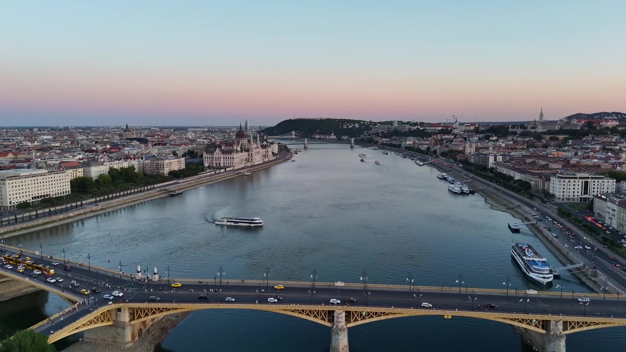Il Parlamento ungherese e il Ponte Margherita a Budapest lungo il Danubio al crepuscolo, con barche, traffico di tram e luci della città nello skyline urbano, ripresi da un drone