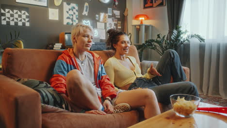 girlfriends relaxing on couch in living room, watching tv and laughing