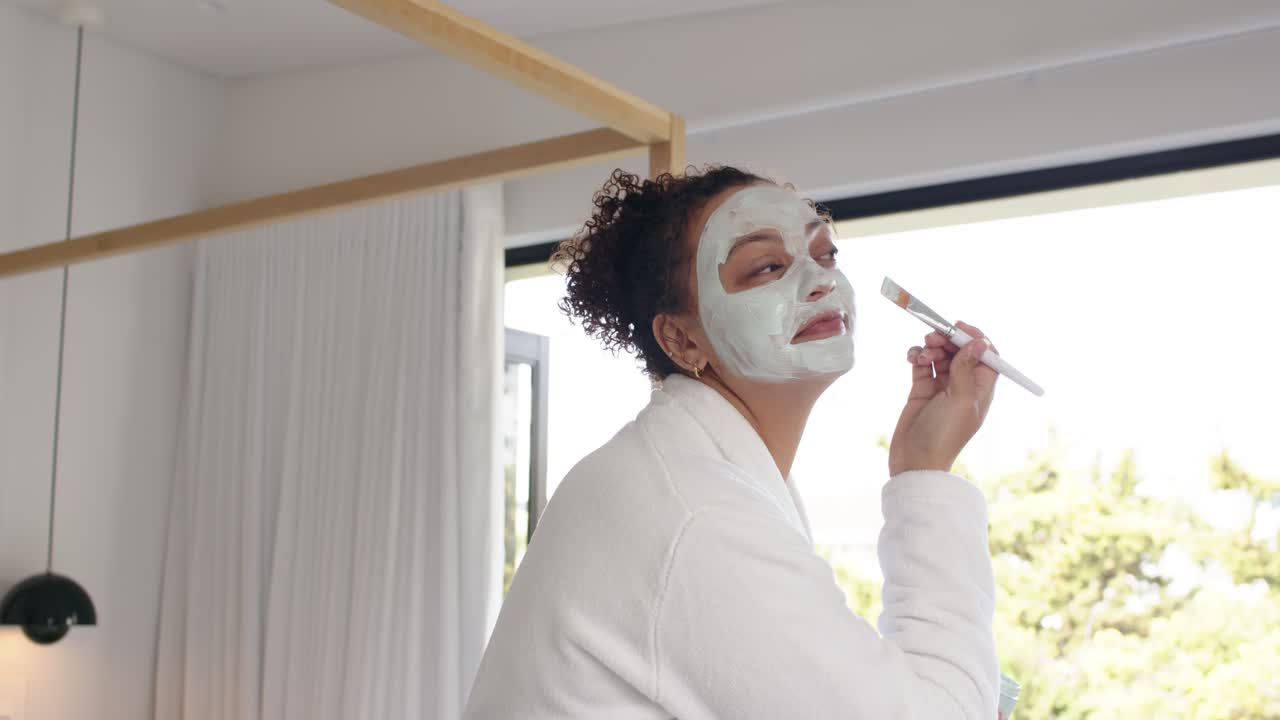 Mujer afroamericana con albornoz aplicándose una mascarilla de menta en casa y mojando el pincel en un tarro para el cuidado de la piel