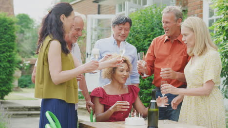 group of mature friends at surprise birthday party for woman in garden with cake and champagne