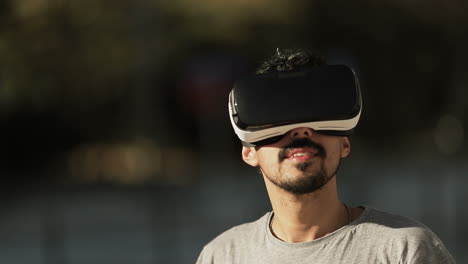 Nahaufnahme-Des-Gesichts-Eines-Jungen-Arabischen-Mannes-Mit-Dunklem-Lockigem-Haar-Und-Bart-Im-Grauen-T-Shirt,-Der-Mit-Einer-Virtual-Reality-Brille-Im-Park-Ist