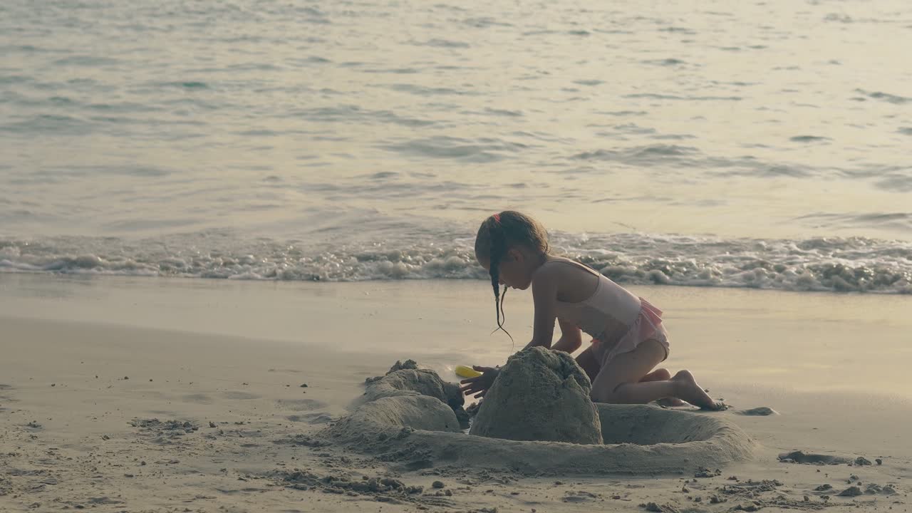 플레이트를 입은 귀여운 소녀는 저녁 해변에 모래 성을 짓습니다.