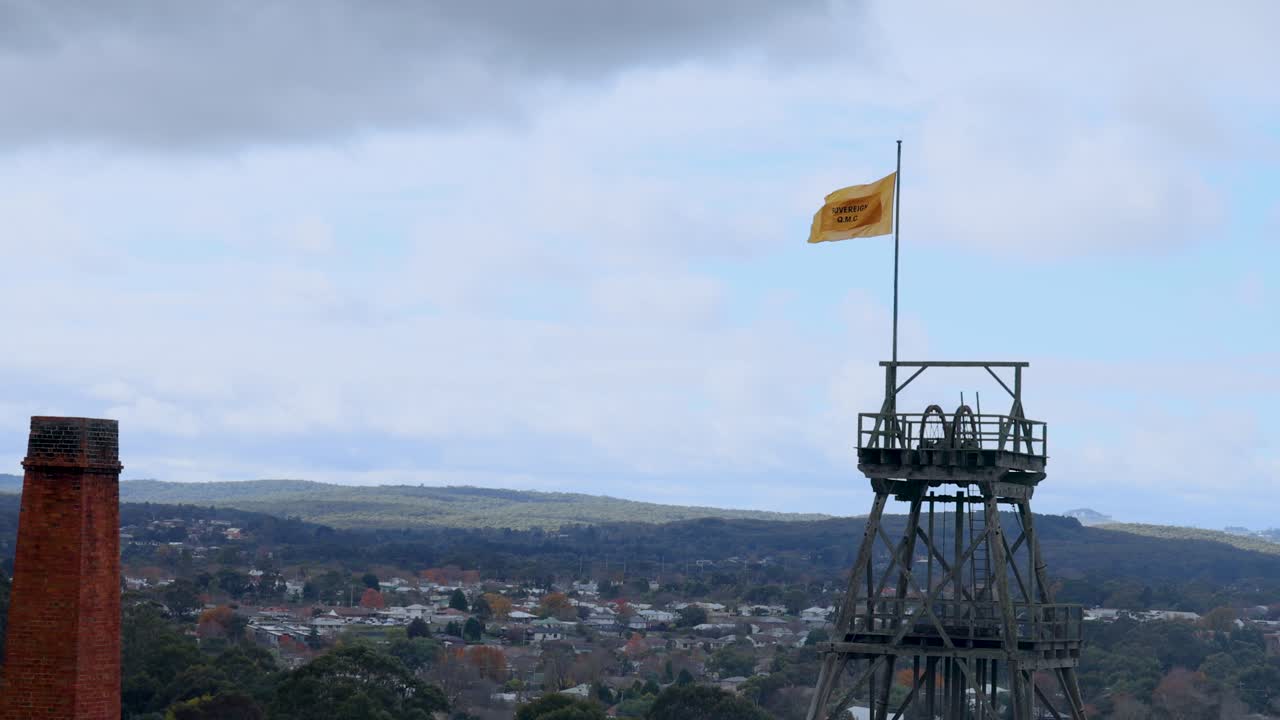 Uma bandeira tremula no topo de uma torre contra o horizonte nublado de Melbourne. O vídeo captura o movimento dinâmico da bandeira.