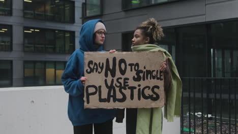 Zwei-Junge-Klimaaktivistinnen-Halten-Ein-Banner-Hoch-Und-Protestieren-Gegen-Einwegkunststoffe,-Während-Sie-Auf-Kamera-1-Blicken