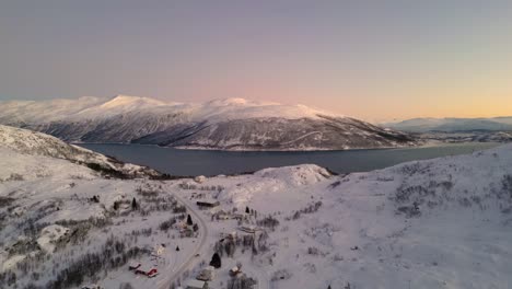 Aerial-drone-shot-over-a-snow-filled-mountain-range-surrounding-a-lake-during-a-sunrise-winter-morning-in-Ersfjordvegen-Norway