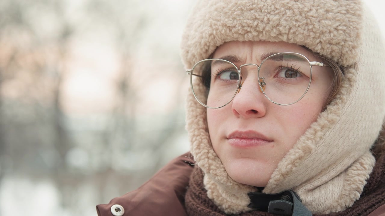 Primo piano all'altezza degli occhi mostra una giovane donna in un inverno nevoso che si sistema gli occhiali con una mano guantata, con una leggera rotazione della testa e un'espressione calma e concentrata, avvolta in un cappello di pile, sciarpa e cappotto, con donna, inverno, neve