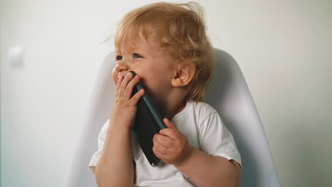 little-boy-holds-cell-phone-near-mouth-sitting-on-chair