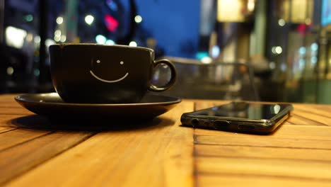 a black coffee cup with a smiley face on it sitting on a table outside at night with a phone next to it